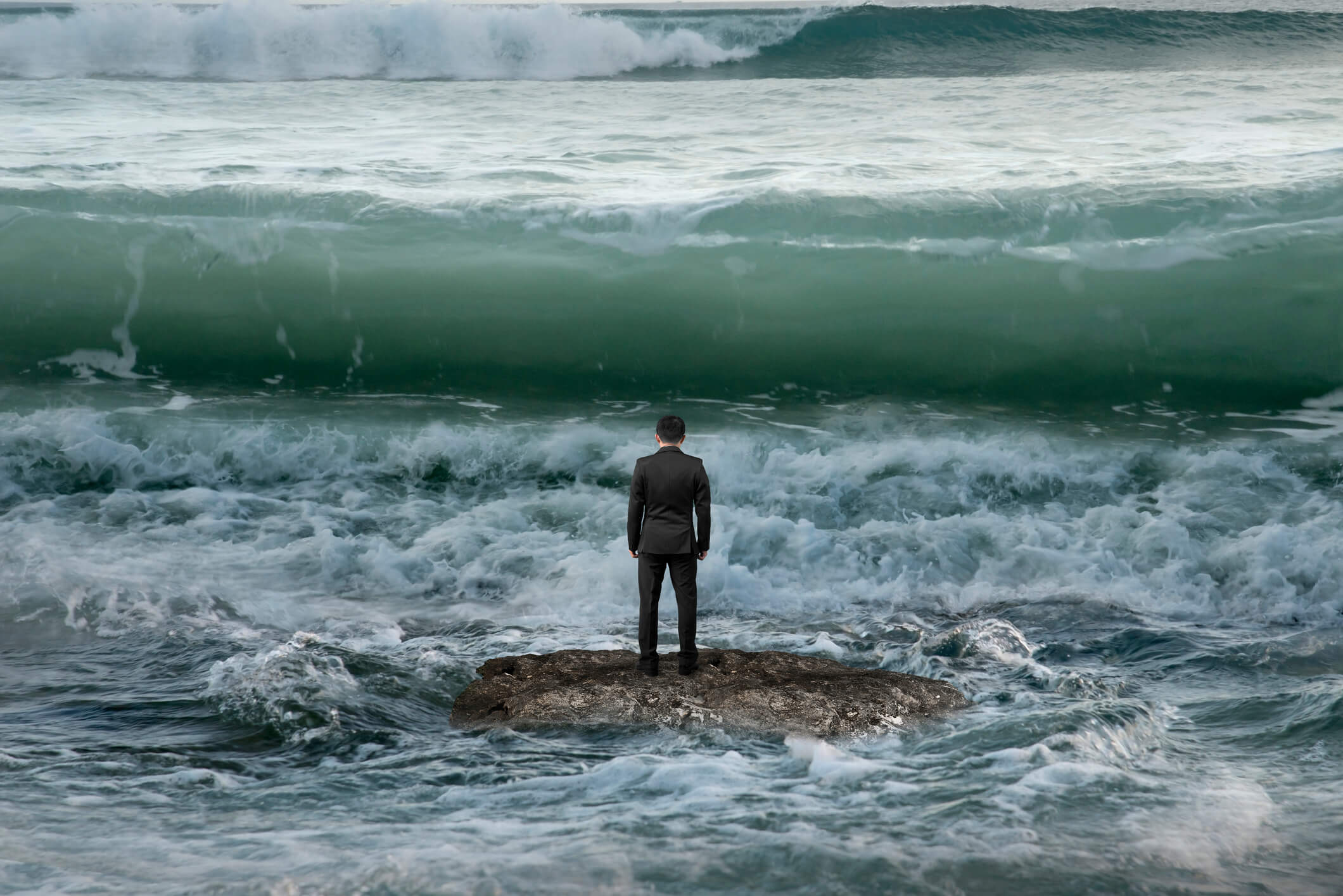 Businessman Standing On Rock In The Ocean Facing Oncoming Waves