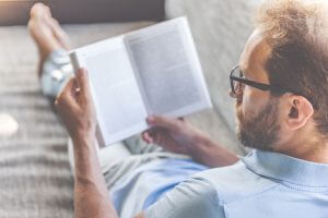 Back view of handsome young businessman in casual clothes and eyeglasses reading a book while lying on couch at home